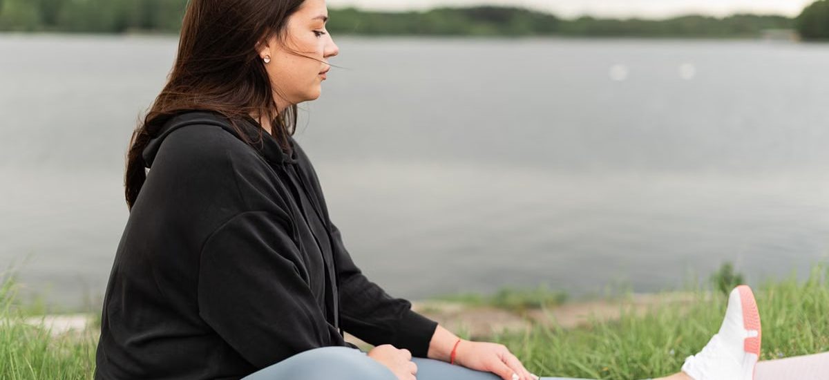 Woman meditating outdoors by a tranquil lakeside, fostering health and wellness.