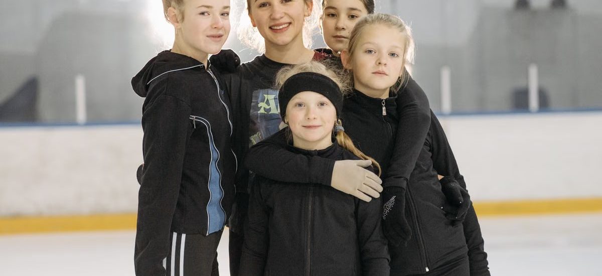 A group of young girls smiling and posing on an indoor ice rink wearing warm winter clothing.