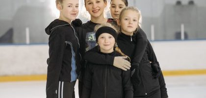 A group of young girls smiling and posing on an indoor ice rink wearing warm winter clothing.