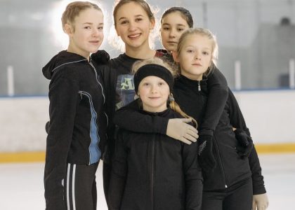 A group of young girls smiling and posing on an indoor ice rink wearing warm winter clothing.