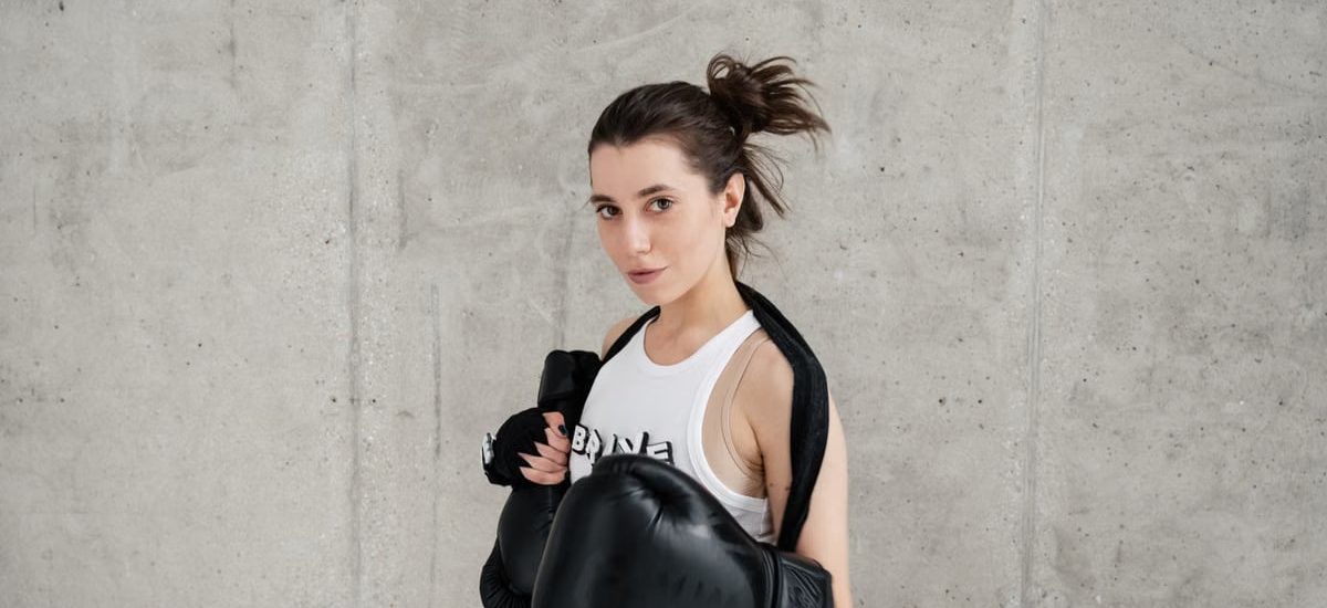 Confident young woman posing with boxing gloves against a concrete wall, showcasing style and sportiness.