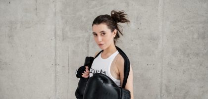 Confident young woman posing with boxing gloves against a concrete wall, showcasing style and sportiness.